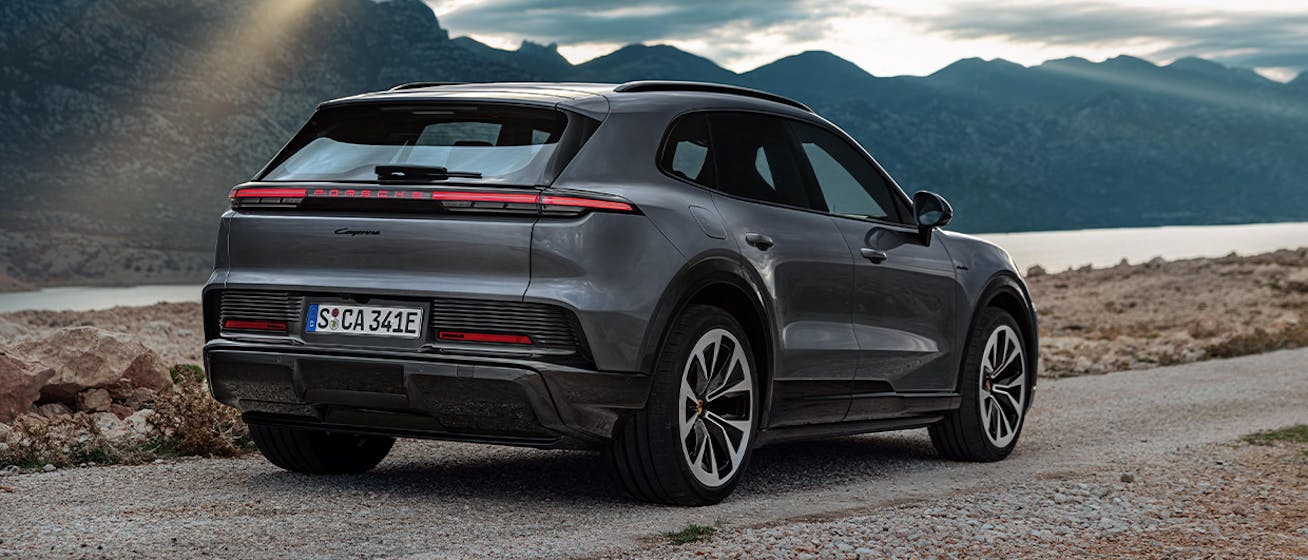 Rear three-quarter view of Porsche Cayenne Electric parked on road by lake, surrounded by mountain lit by shafts of sunlight