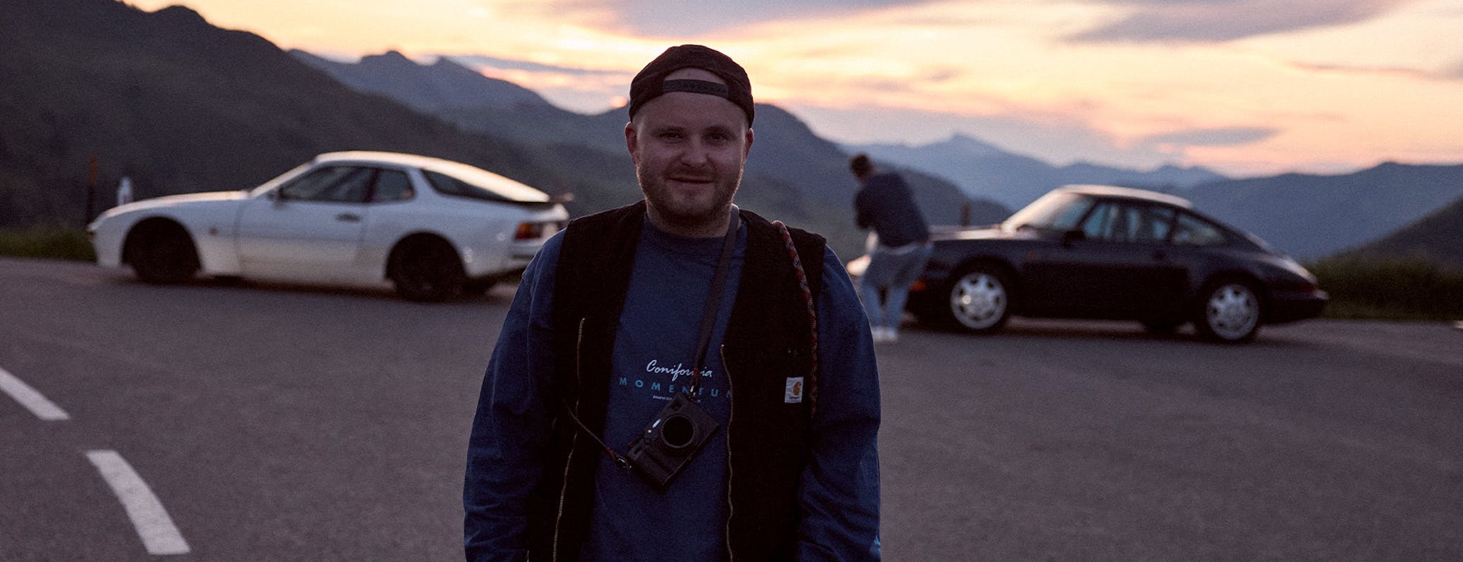 Man with camera at sunset, classic Porsche cars behind him