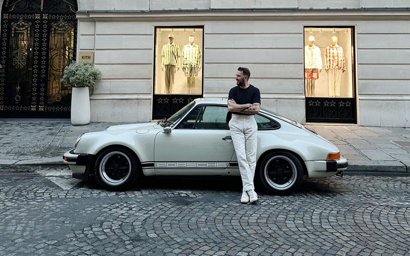 Man standing next to classic Porsche 911 parked on cobbled street outside clothing store in Paris