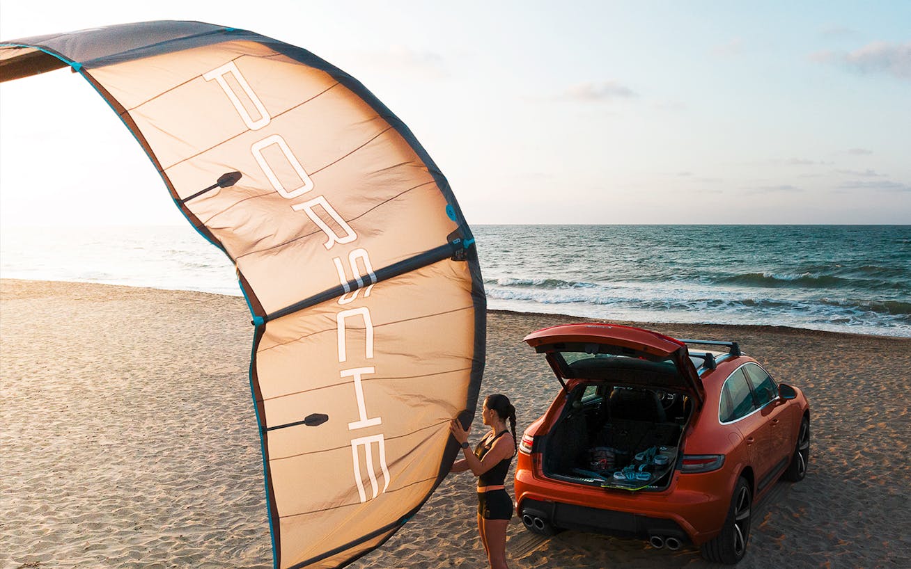 Female kitesurfer holds onto sail, Porsche Cayenne and ocean behind