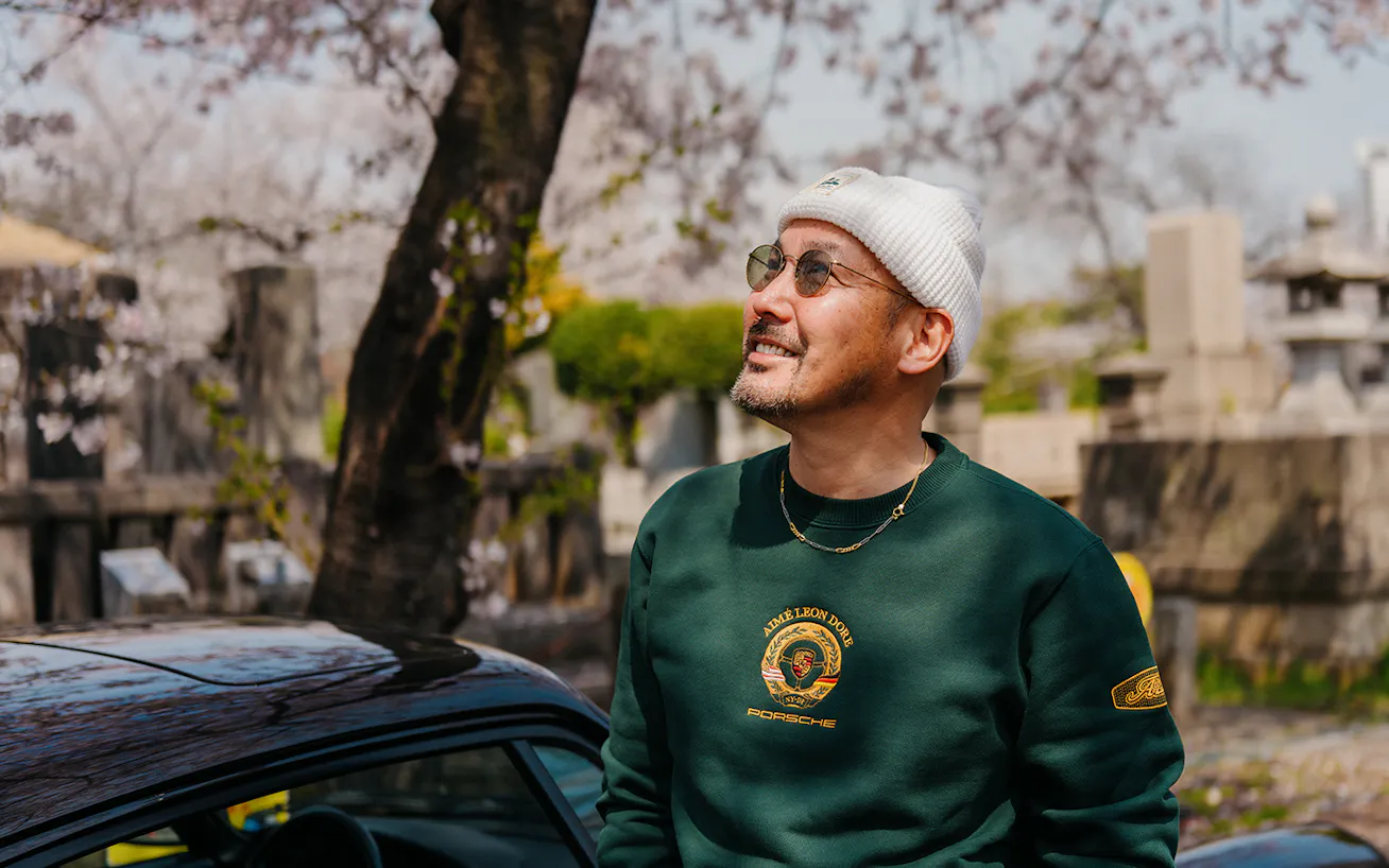 Man in sunglasses looking up at cherry blossom trees in Japan, classic Porsche 911 S/C behind