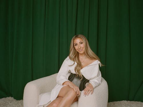 Hanna Lux Davis sits in a white armchair against a dark green background and smiles