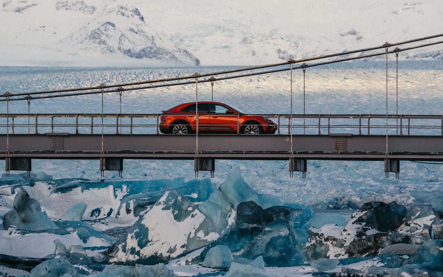 Wide shot of Porsche Macan Electric driving on a bridge over a frozen lagoon in Iceland with snow-covered mountains in the background