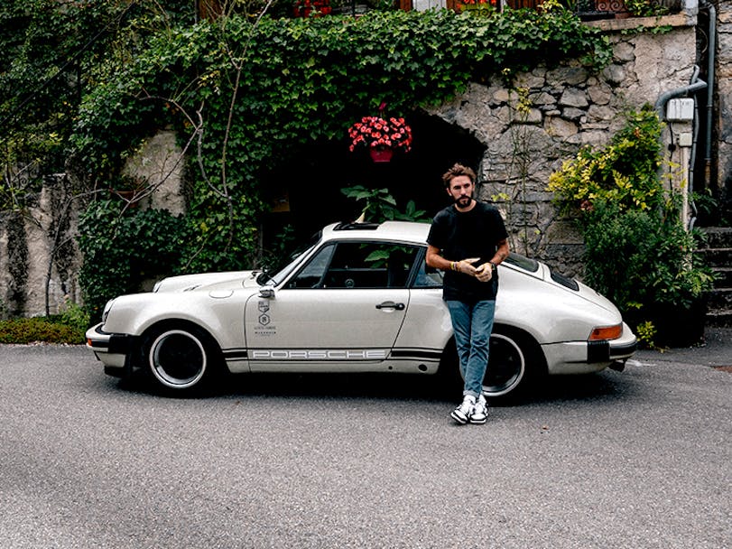 Man leaning on cream coloured 1987 Porsche 911 outside French stone built restaurant thumbnail