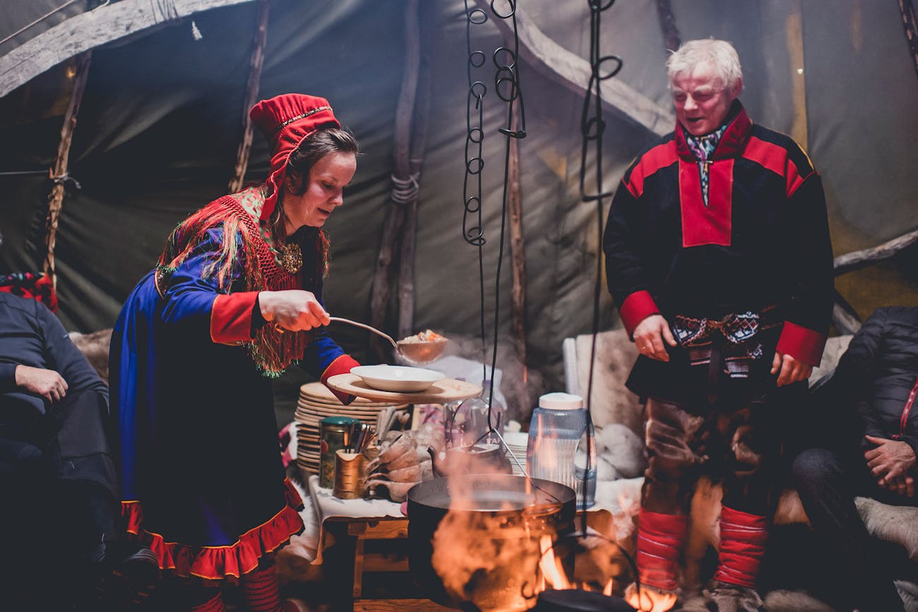 Sami woman ladles soup from pot over the tent fireplace