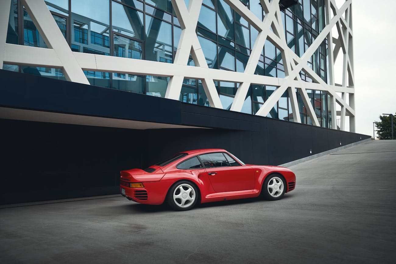 Red Porsche 959 production car driving past modern office building