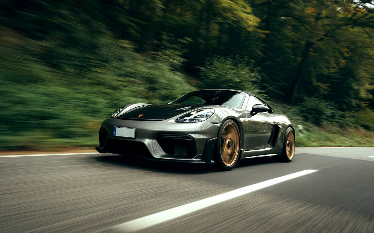 Grey Porsche 718 Spyder RS driving on road, trees behind