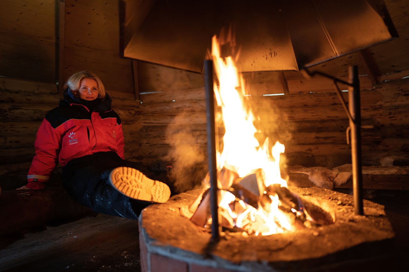 A woman warming herself by an open fire in a hut on the Porsche Ice Experience accompanying programme in Levi, Finland