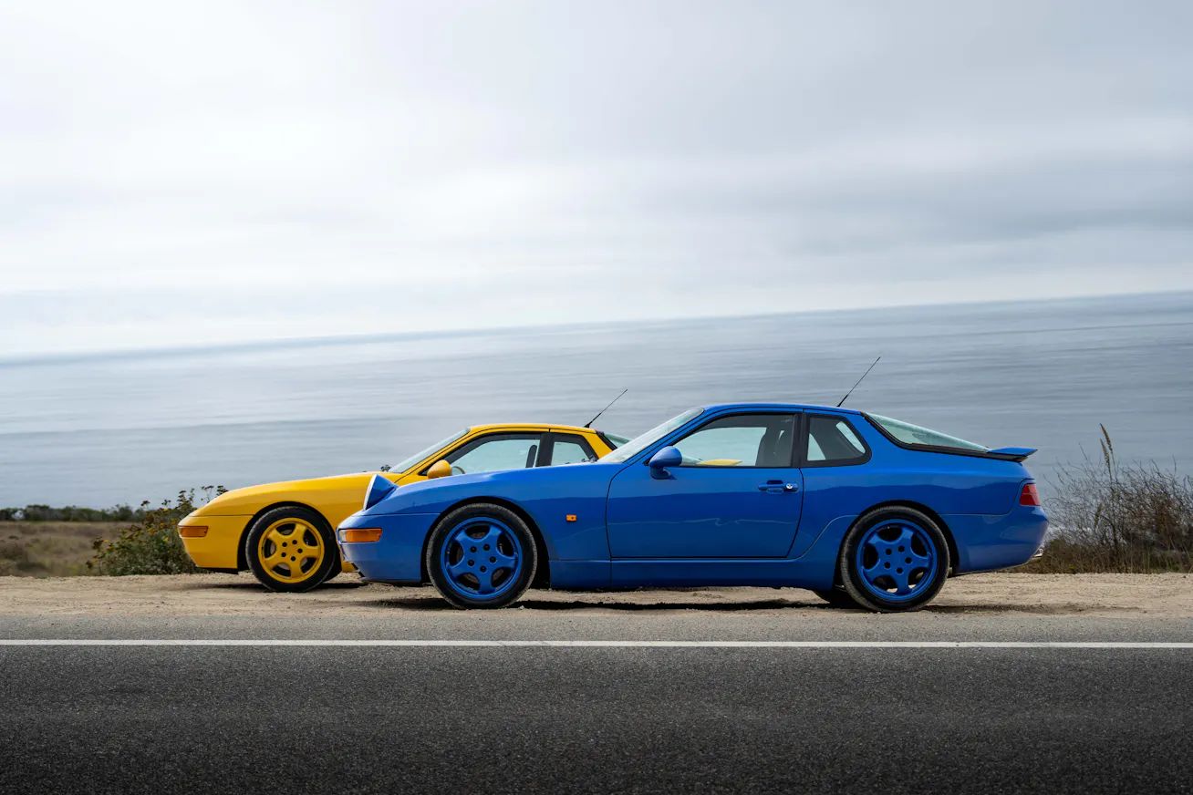 Side profiles of two Porsche 968 Club Sport cars in Speed Yellow and Maritime Blue with backdrop of Pacific Ocean at Big Sur, California