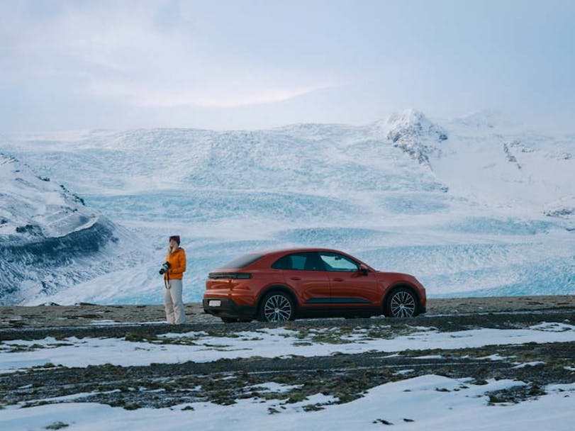 Woman standing behind Porsche Macan Electric in Iceland with ice-covered hills in the background 