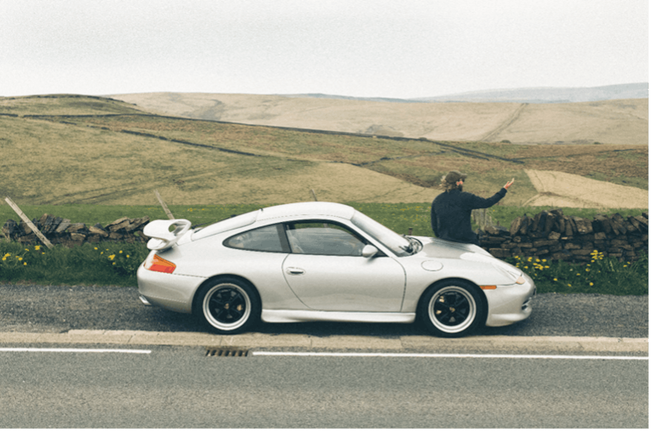 Man sits on bonnet of Porsche 911 (996) overlooking countryside