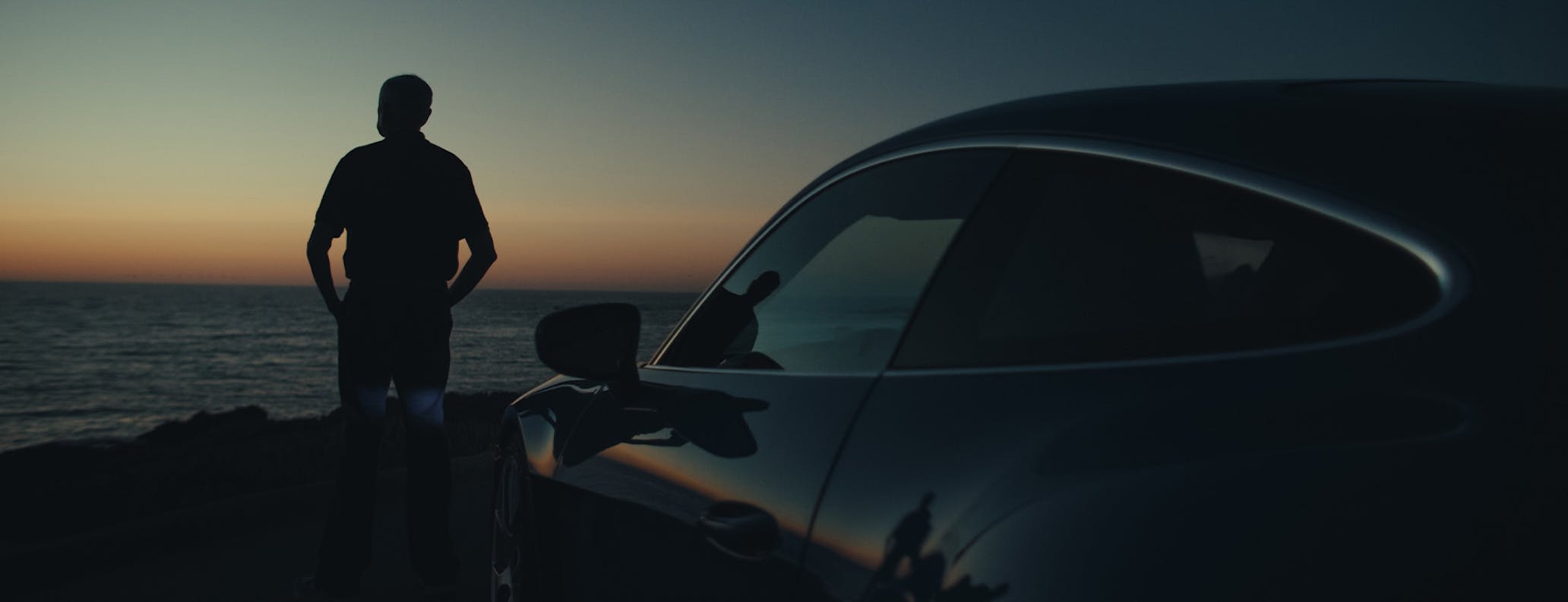 Profile of man by sea with Porsche sportscar behind him