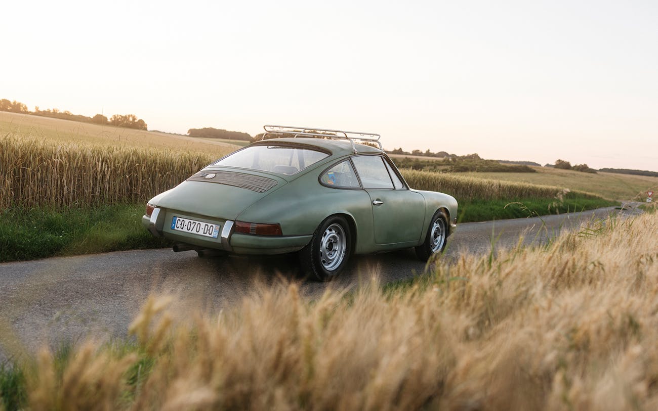 Green Porsche 912 driving on country road surrounded by fields