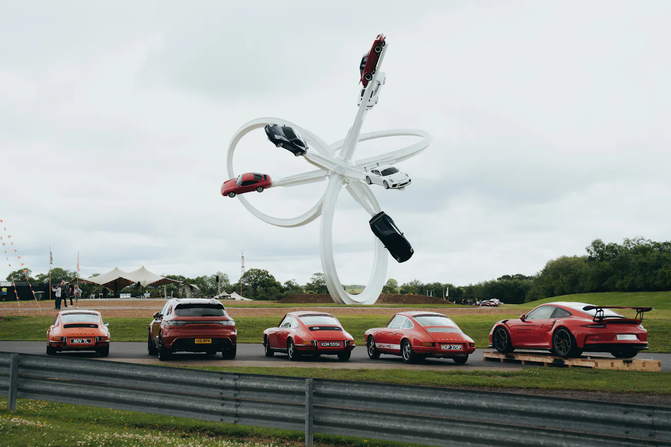 Aerial sculpture showing five Porsche models with a row of Porsche cars in the foreground