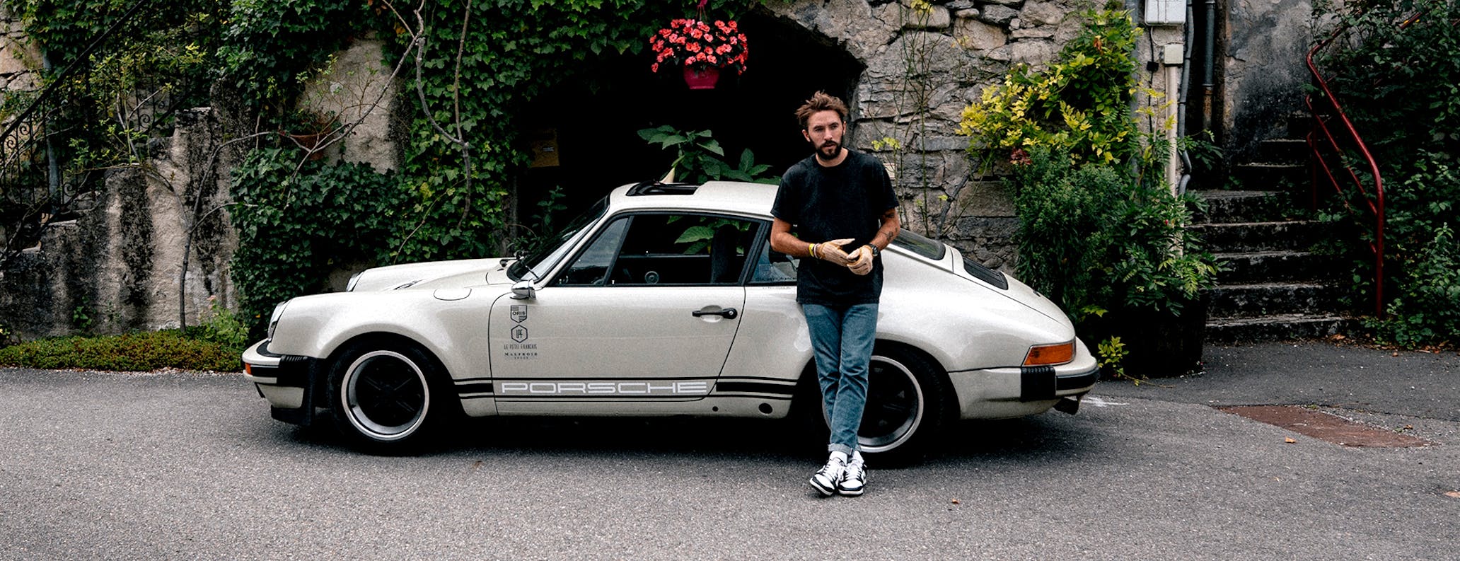 Man leaning on cream coloured 1987 Porsche 911 outside French stone built restaurant desktop