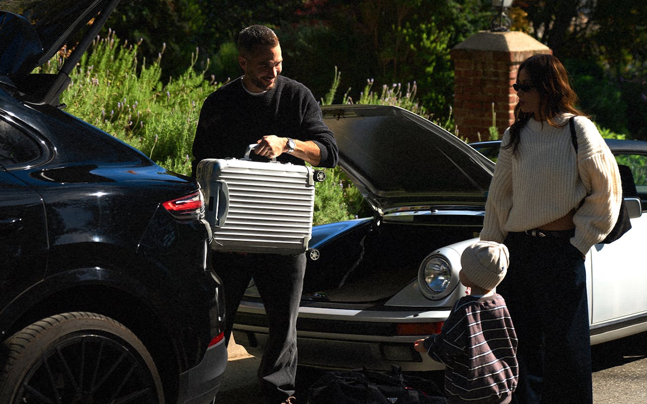 Julie Sariñana, aka Sincerely Jules, with husband, loading luggage into classic Porsche 911 Targa and Porsche Cayenne coupé (E3)