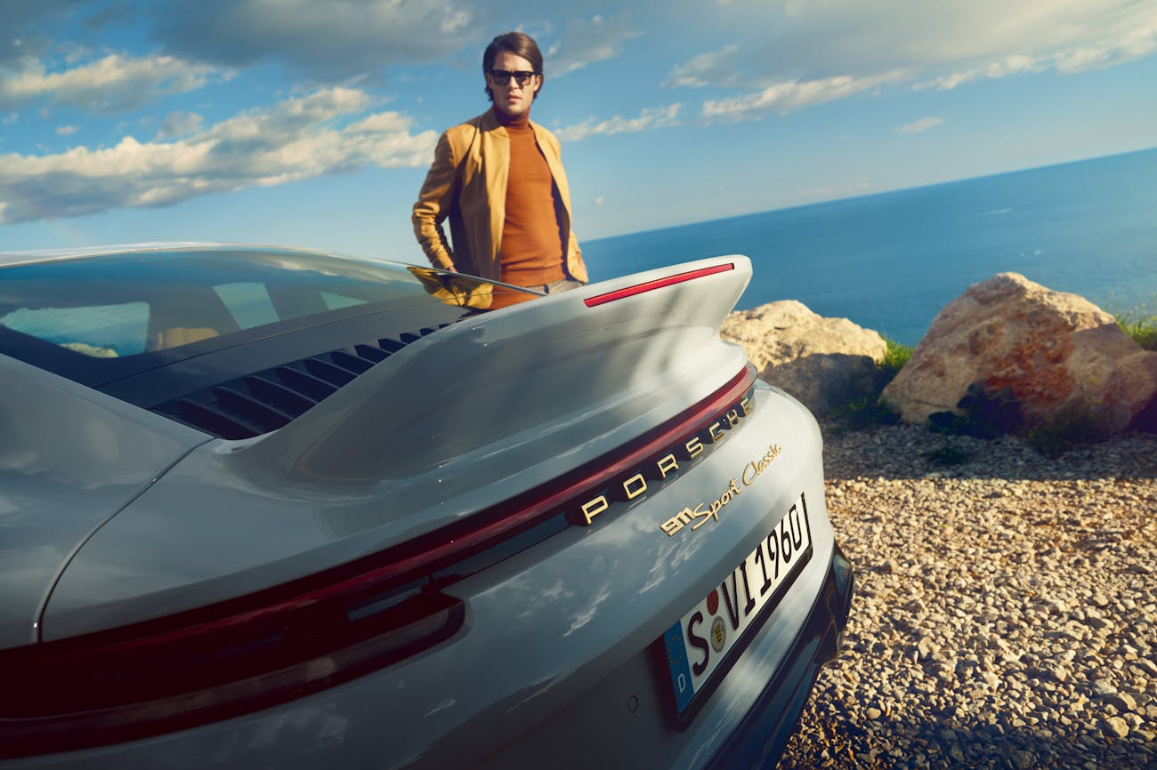 Man standing behind rear of grey Porsche with ducktail, ocean in background
