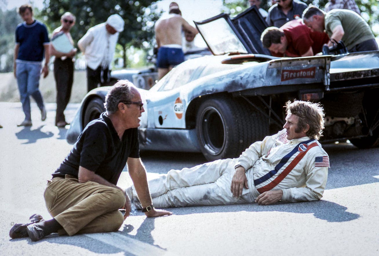 Director of the movie Le Mans, John Sturges, sits on the floor talking to actor Steve McQueen, dressed in a racing suit with Porsche 917 K race car behind them