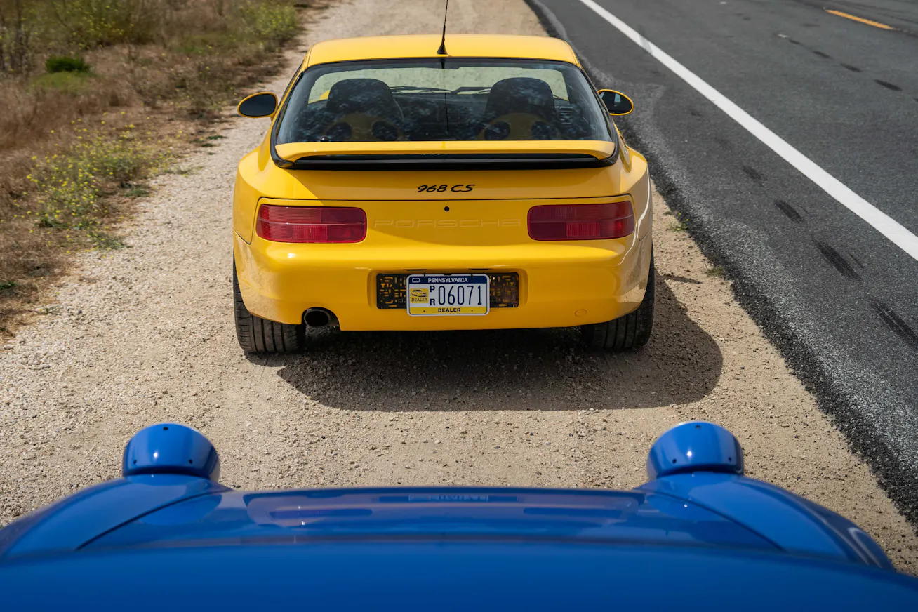 Rear of Porsche 968 Club Sport in Speed Yellow and nose of Porsche 968 Club Sport in Maritime Blue besides the Pacific Coast Highway, Big Sur, California