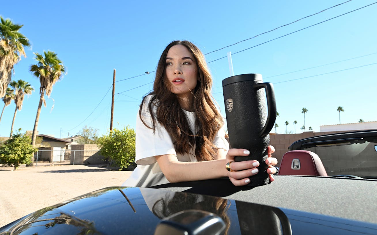 Woman holding thermos cup, leaning on Porsche car