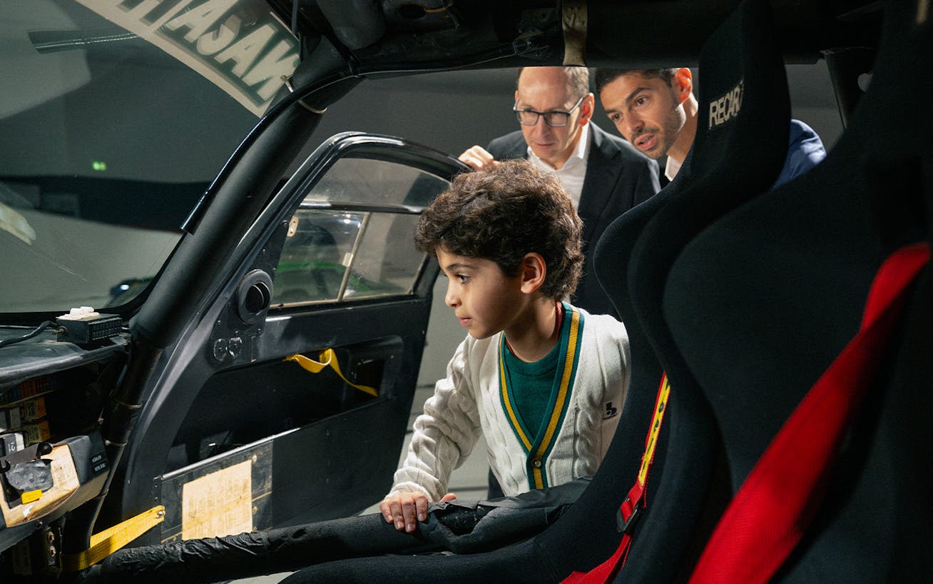 Child explores car interior while two men observe closely