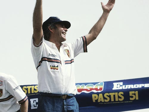 René Metge is standing at the finish line and celebrating with his hands in the air.