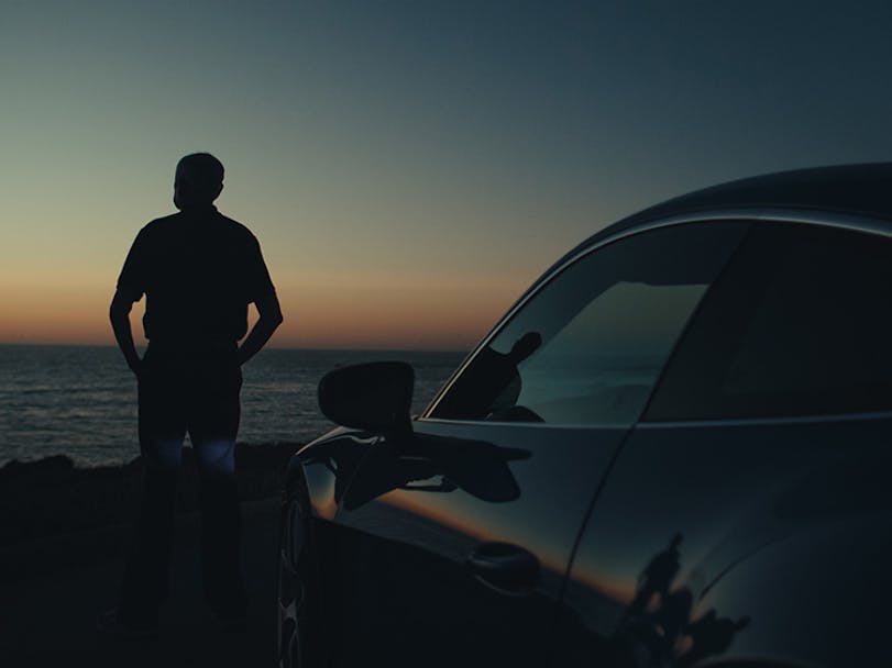 Profile of man by sea with Porsche sportscar behind him